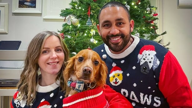 a man and woman pose with their dog all dressed in Christmas jumpers in front of a Christmas tree