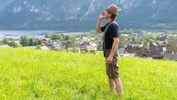 A man in traditional Austrian clothing stands in a grassy field overlooking a picturesque village nestled between mountains on a sunny day. Shouting into the distance.