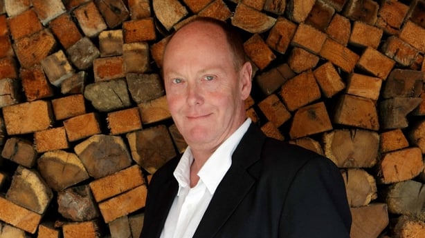 Paul McCreesh stands in front of a big pile of wooden blocks, smiling at the camera. 