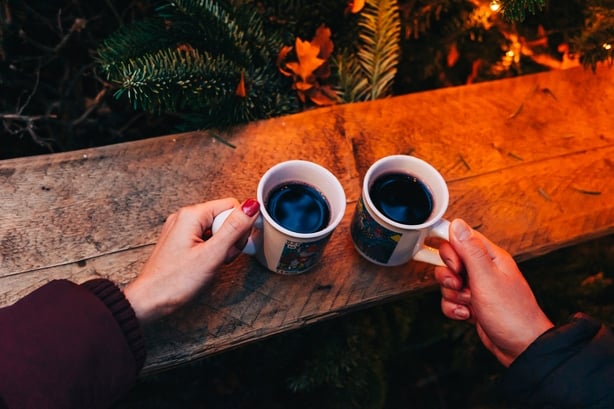 Two people clink black mugs filled with glühwein (mulled wine)in a glowing, festive market scene. Holiday lights, cozy textures, and seasonal cheer create a joyful, together moment.