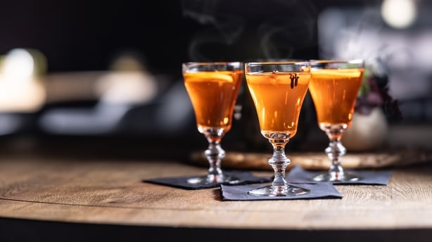 On a dark wooden table are three stemmed glasses with steam rising from hot Aperol, decorated with a slice of orange and cloves.