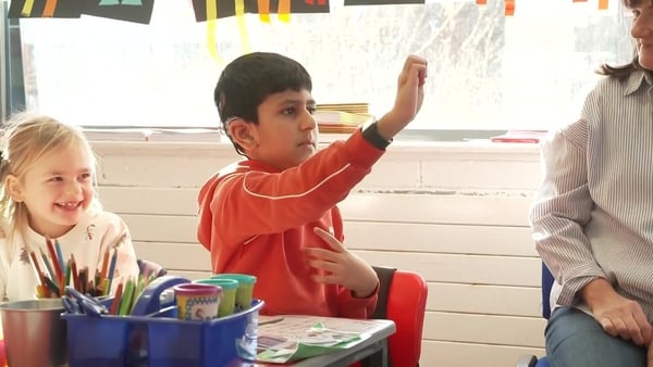 A boy sits at a desk using Irish Sign Language in a classroom