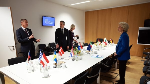 A group of people including Ursula von der Leyen stand around a table with various small flags and coffee cups