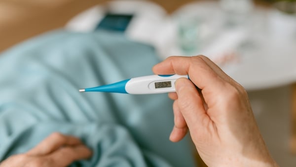 Over-the-shoulder view of an unrecognizable woman looking at the thermometer because she is sick while sitting on the sofa covered with a blanket.