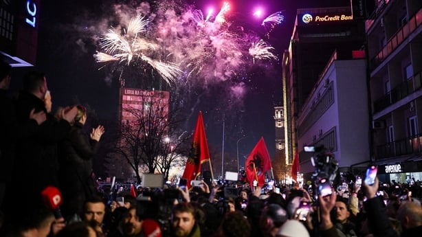 Hundreds of people gather on a street celebrating. There are fireworks in the sky above.