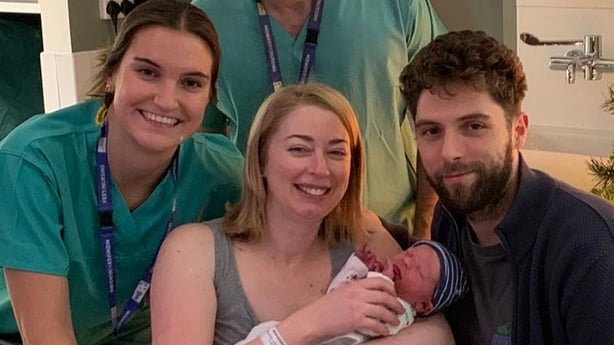 three people are pictured with a fourth in the background and a newborn baby with a blue striped hat