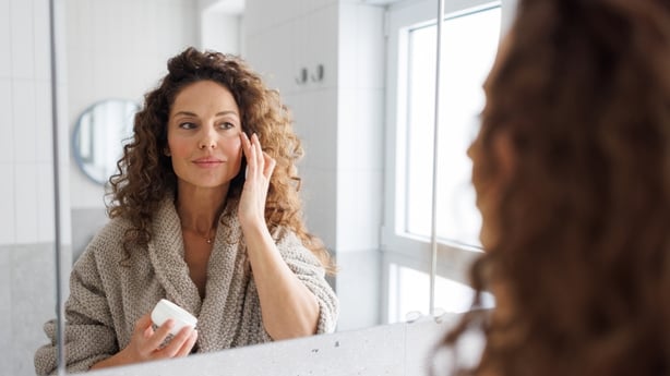woman applies face cream to her face in a bright, modern bathroom