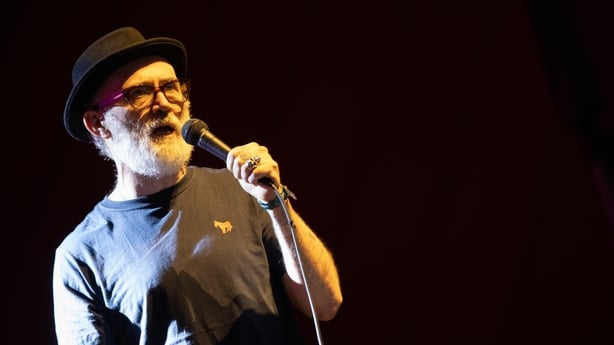 Tommy Tiernan wears glasses and a hat, and holds a mic while performing on stage