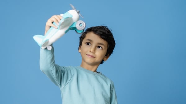 Cheerful young boy holding up a white and blue toy airplane, looking at it with wonder and a smile. Studio portrait shot against a solid blue background