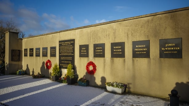 A yellow wall with a dozen black plaques can be seen against a bright blue sky.