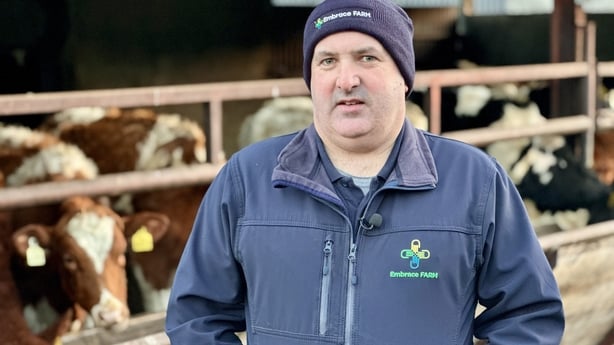 A man wearing a navy jacket and hat stands in front of barn containing several cows.