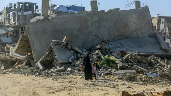 a woman and two small children walk on dusty roads with destroyed buildings behind them
