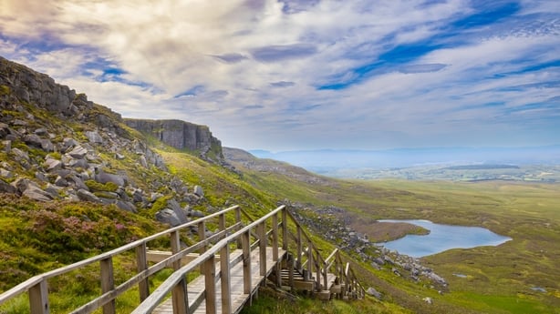 Cuilcagh Mountain boardwalk trail, known as Stairway to Heaven, in County Fermanagh, Northern Ireland, featuring wooden steps, rocky hills, scenic moorland, and a lake under a cloudy sky.