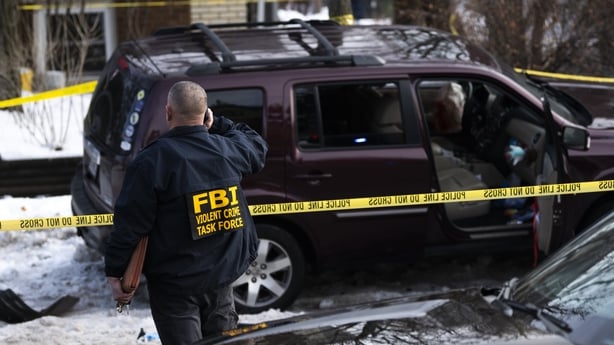 A man wearing a jacket with FBI written on it stands on front of a crime scene tape and a maroon coloured vehicle