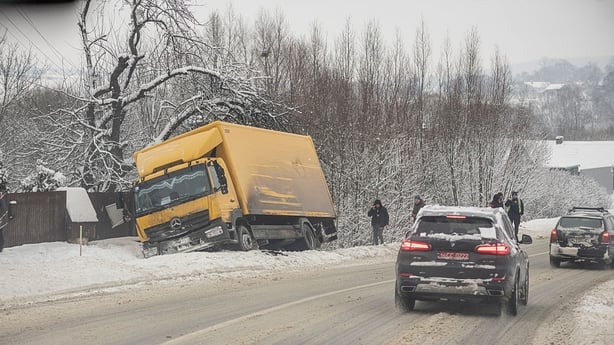 Cars drive along a snow-covered road past a yellow truck that is stuck on the roadside 