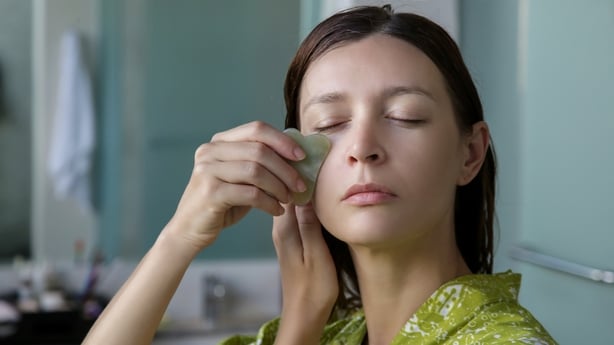 Young woman is doing Gua Sha facial massage in bathroom in front of the mirror