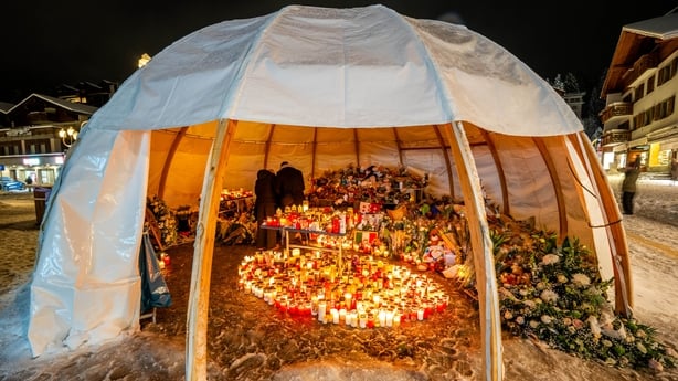 Attendees gather in mourning, placing messages, candles, and flowers at a memorial site in front of the bar 