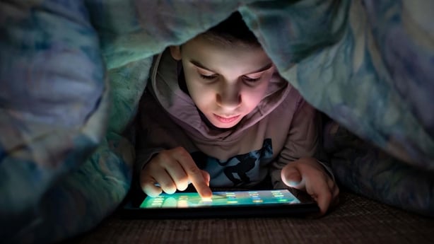 A young boy playing on a tablet under a duvet at night