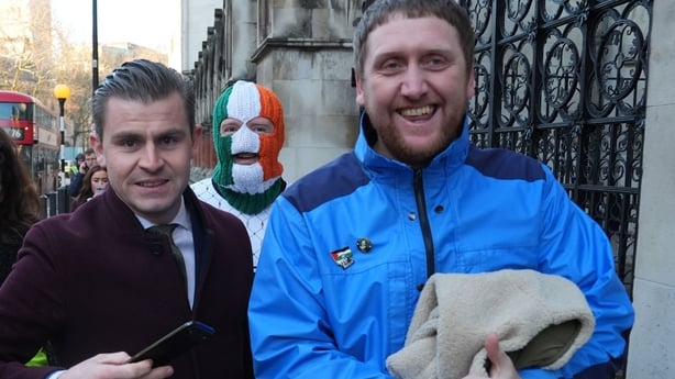 Three men are photographed with one wearing a green white and gold balaclava covering his face
