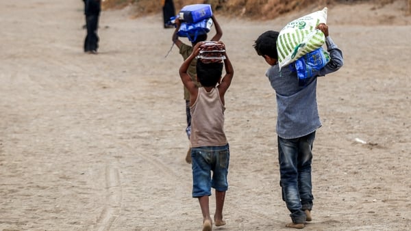 Two young children walk away from the camera down a dirt track. They are carrying large bags of aid over their shoulders.