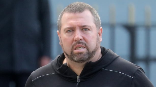 A close up head and shoulders image of a man with brown hair and brown stubble wearing a dark coloured jumper.