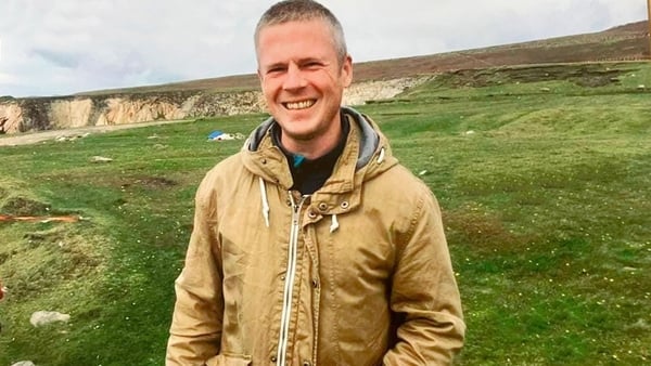 A smiling man stands in a green field. He is wearing a yellow jacket.