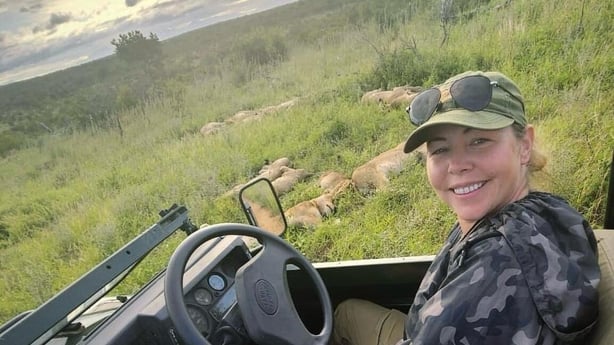 A woman sits behind the wheel of a jeep in South Africa, with lions lying in the grass behind her