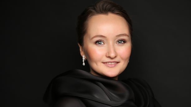 Julia Lezhneva smiles directly at the camera, wearing a black scarf with a black blackground behind her