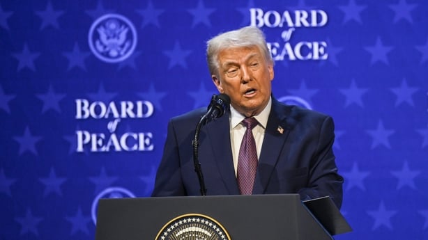 Donald Trump wears a navy suit, white shirt and purple tie. He is standing at a podium with a microphone. The backdrop is blue and reads: 'Board of Peace'.