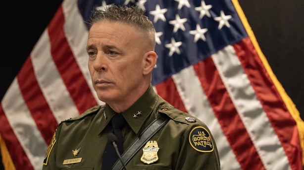 Greg Bovino stands in front of an American flag.