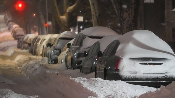 Dozens of parked cars along the side of a street are covered in snow.