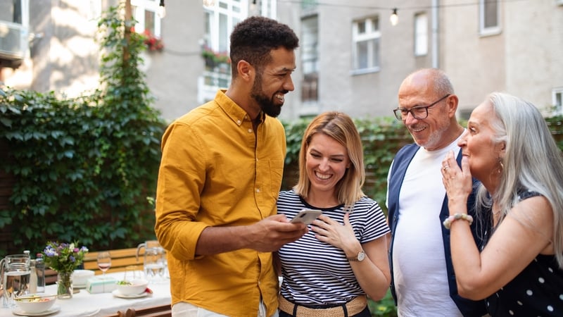 Happy multiracial couple with senior parents standing and using mobile after family dinner outdoors in front or back yard.