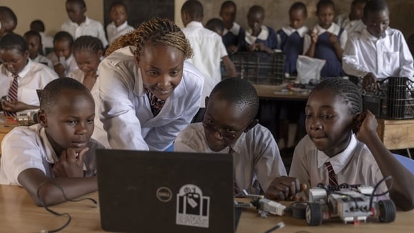 A woman leans over three young children as they work on a laptop