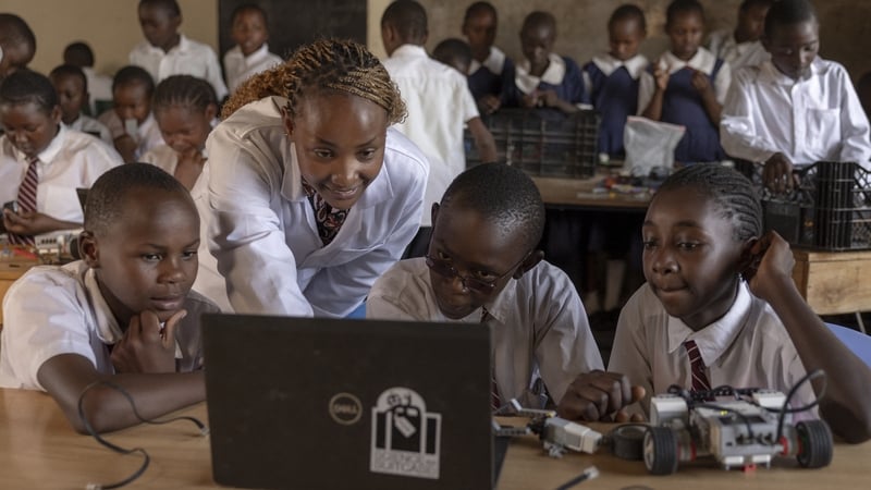 A woman leans over three young children as they work on a laptop