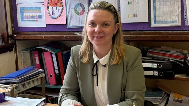 A woman sits at a desk in classroom with posters pinned to a board behind her