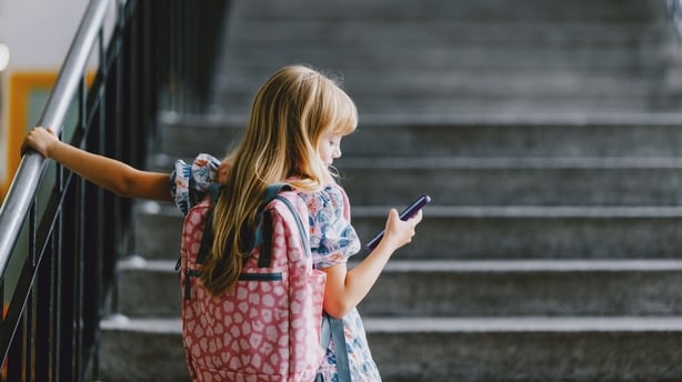 A young girl with a backpack holds her phone while standing on a school staircase. 