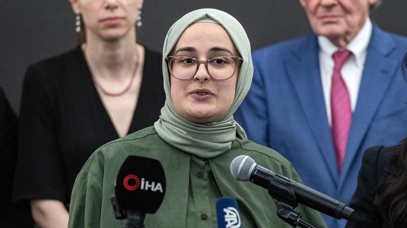 Rumeysa OzturK stands at a podium with two microphones as she addresses a press conference.