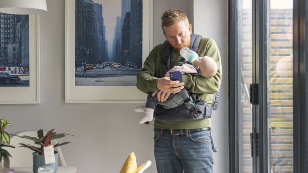 a man feeds a baby while looking at a mobile phone