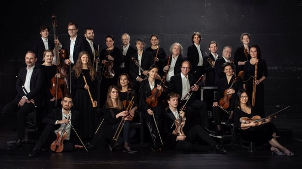 A wide shot of a group of 23 musicians in formal wear, posing with their instruments, with a black background