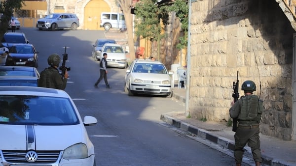 Soldiers wearing camouflage clothing and carrying guns can be seen walking along a street lined with cars.