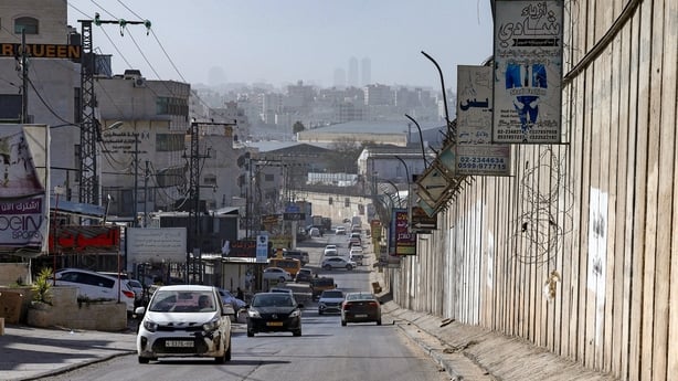 Vehicles can be seen driving along a road. A high fence with barbed wire is on one side of the road, while dilapidated buildings are on the other side.