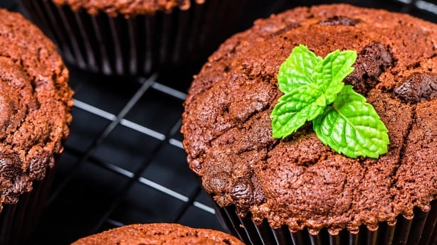 Close-up of rich chocolate muffins, some with chocolate chips, topped with vibrant green mint leaves on a black wire cooling rack.