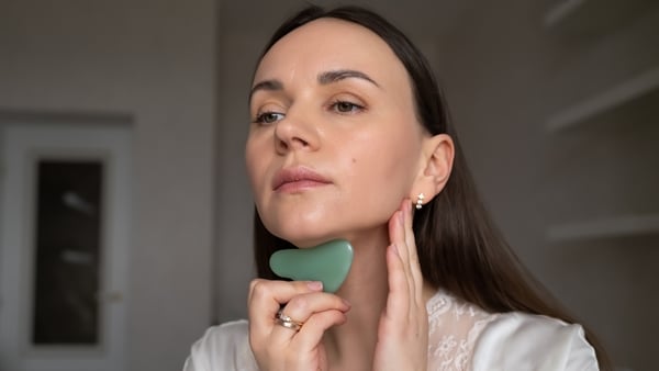 Young woman in silk robe doing facial massage with gua sha scraper while looking in mirror. Home skin care and aesthetic procedures