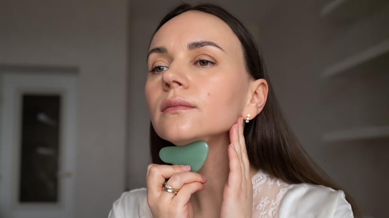 Young woman in silk robe doing facial massage with gua sha scraper while looking in mirror. Home skin care and aesthetic procedures