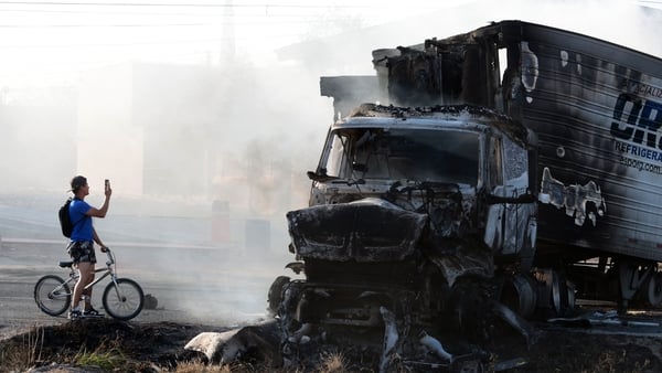 A man riding a bicycle takes a photo of a burned truck, allegedly set on fire by organised crime groups in response to an operation to arrest a high-priority security target