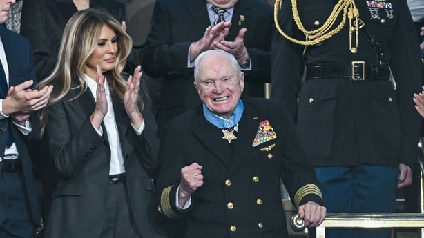 An elderly man smiles and gestures as other people look on and applaud