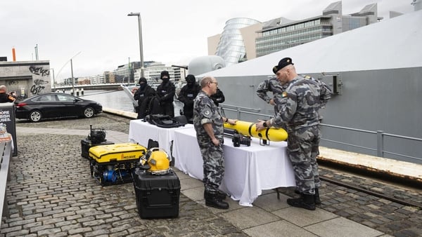 Men in military uniform with military equipment on display with a ship docked behind them