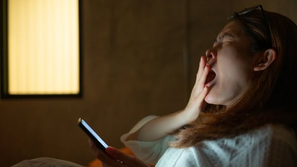 Woman yawning while in bed, holding a phone late at night, only light coming from phone and lamp in background (Photo: Getty Images)