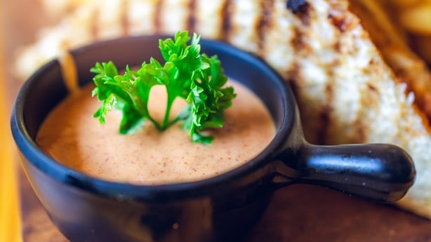 Macro shot of creamy dipping sauce served in a black bowl, topped with a sprig of parsley for freshness. Styled with blurred sandwich and fries in the background, highlighting gourmet food presentation.