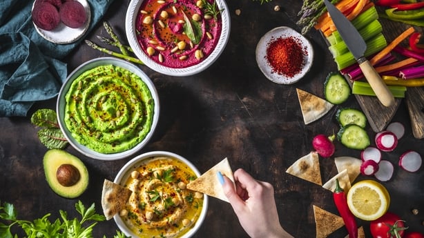Hummus bowls of chickpeas, avocado and beetroot female hand dip with cut vegetables for dipping on dark wooden table background